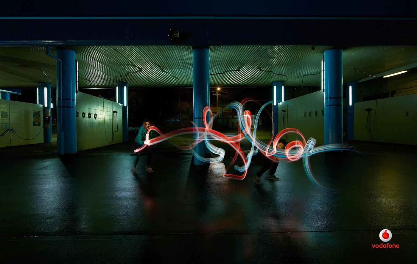 Two people creating light painting trails with handheld light sources in an illuminated covered car park at night.