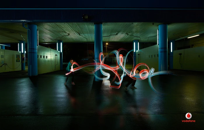 Two people creating light painting trails with handheld light sources in an illuminated covered car park at night.