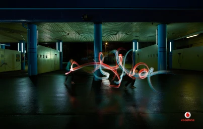 Two people creating light painting trails with handheld light sources in an illuminated covered car park at night.