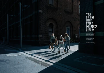 Family of four walking together on a city street in Sydney under strong sunlight cast against a dark brick building exterior.