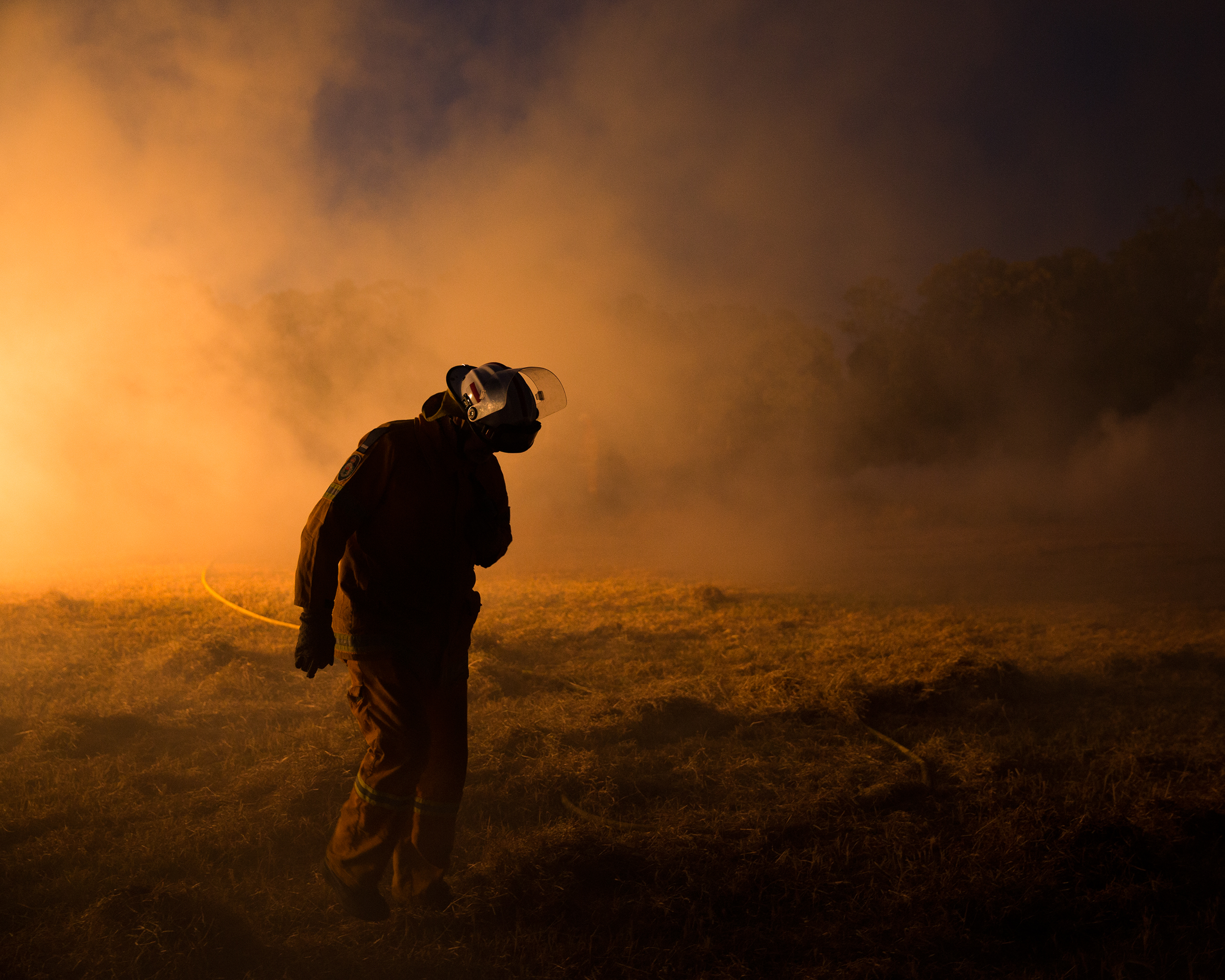 Firefighter in protective gear walks through a smoky field at night, illuminated by orange light. Emergency response scene with obscured visibility.