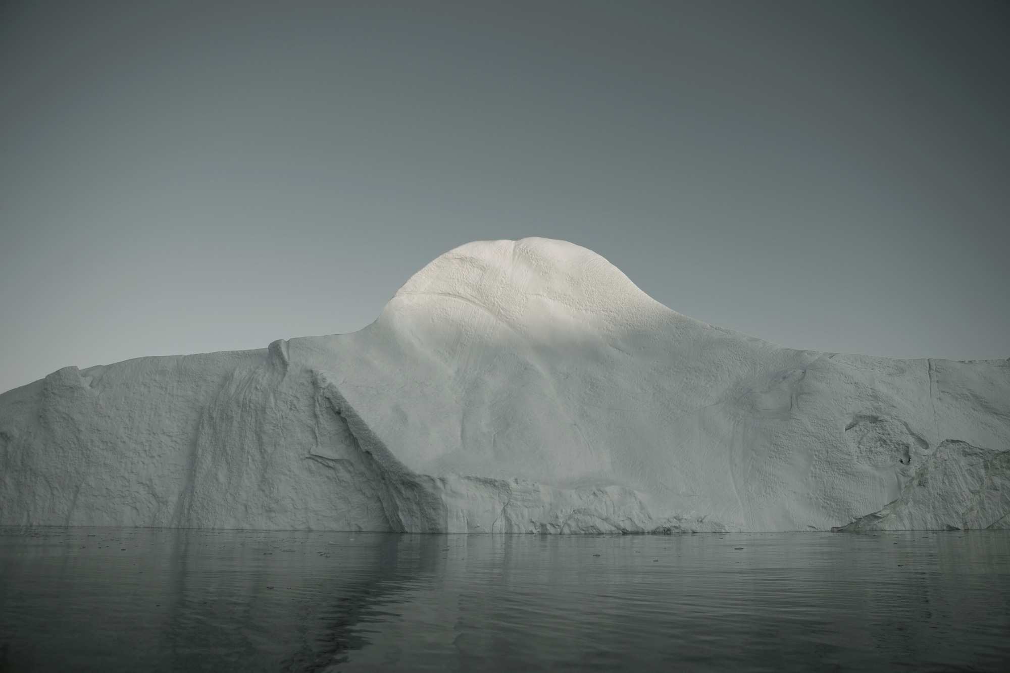 Iceberg floating on the water on a bright day. White and blue glacier sits in the ocean, beneath a gray, cloudless sky.