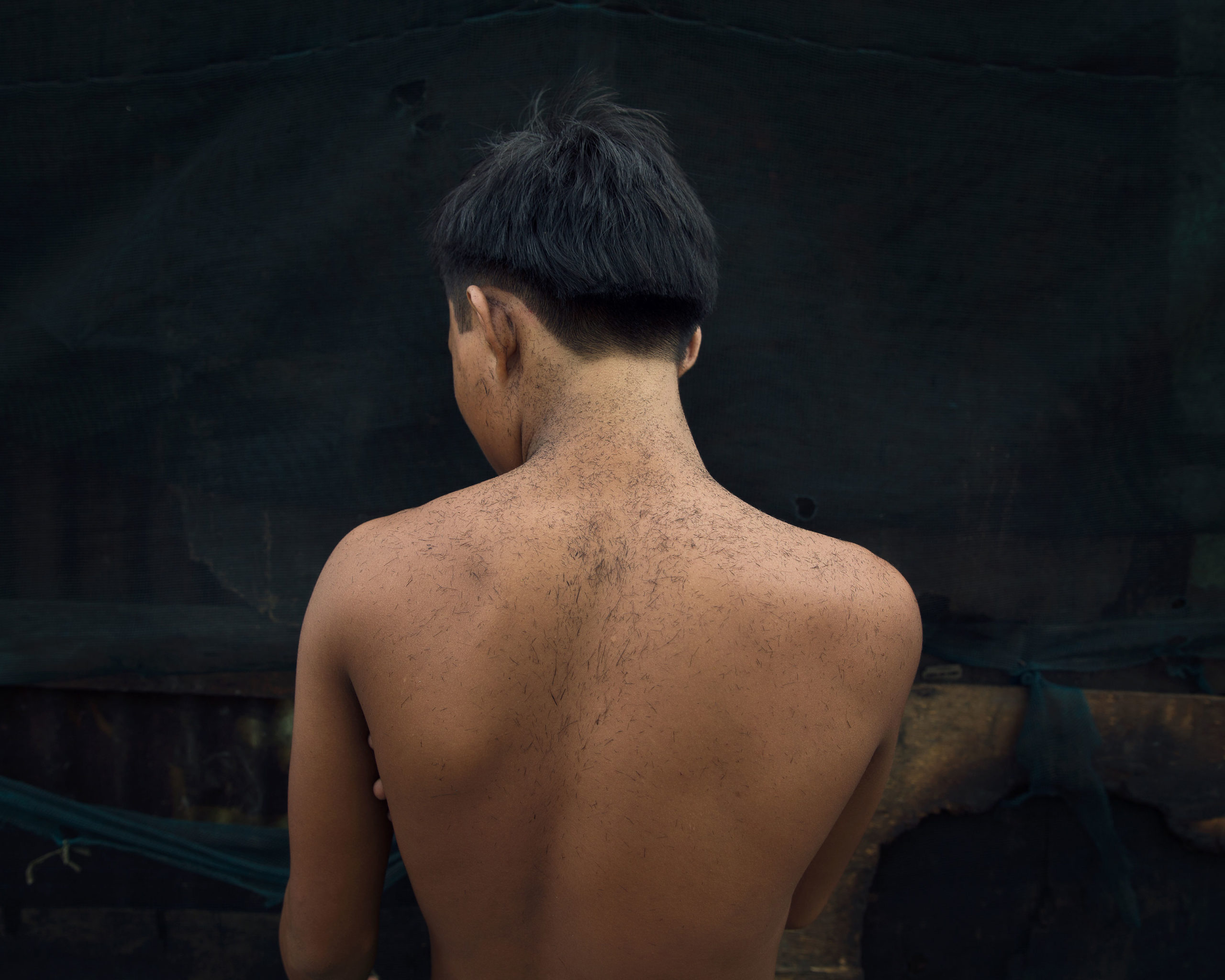A young Asian man stands with his back to the camera, showing a fresh haircut and dark hair against a dark background with wood accents.