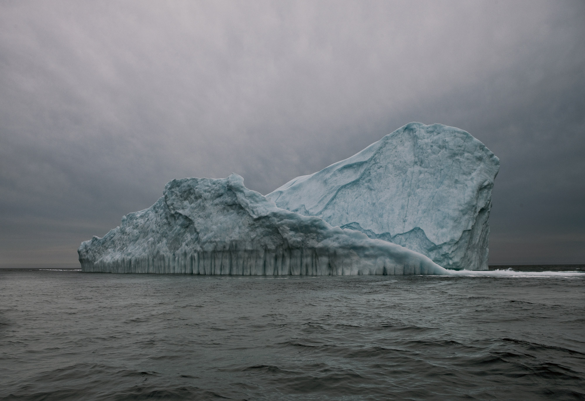 Dramatic wide shot of an iceberg in the ocean, set against a cloudy sky, highlighting its scale and the vastness of the Arctic landscape.