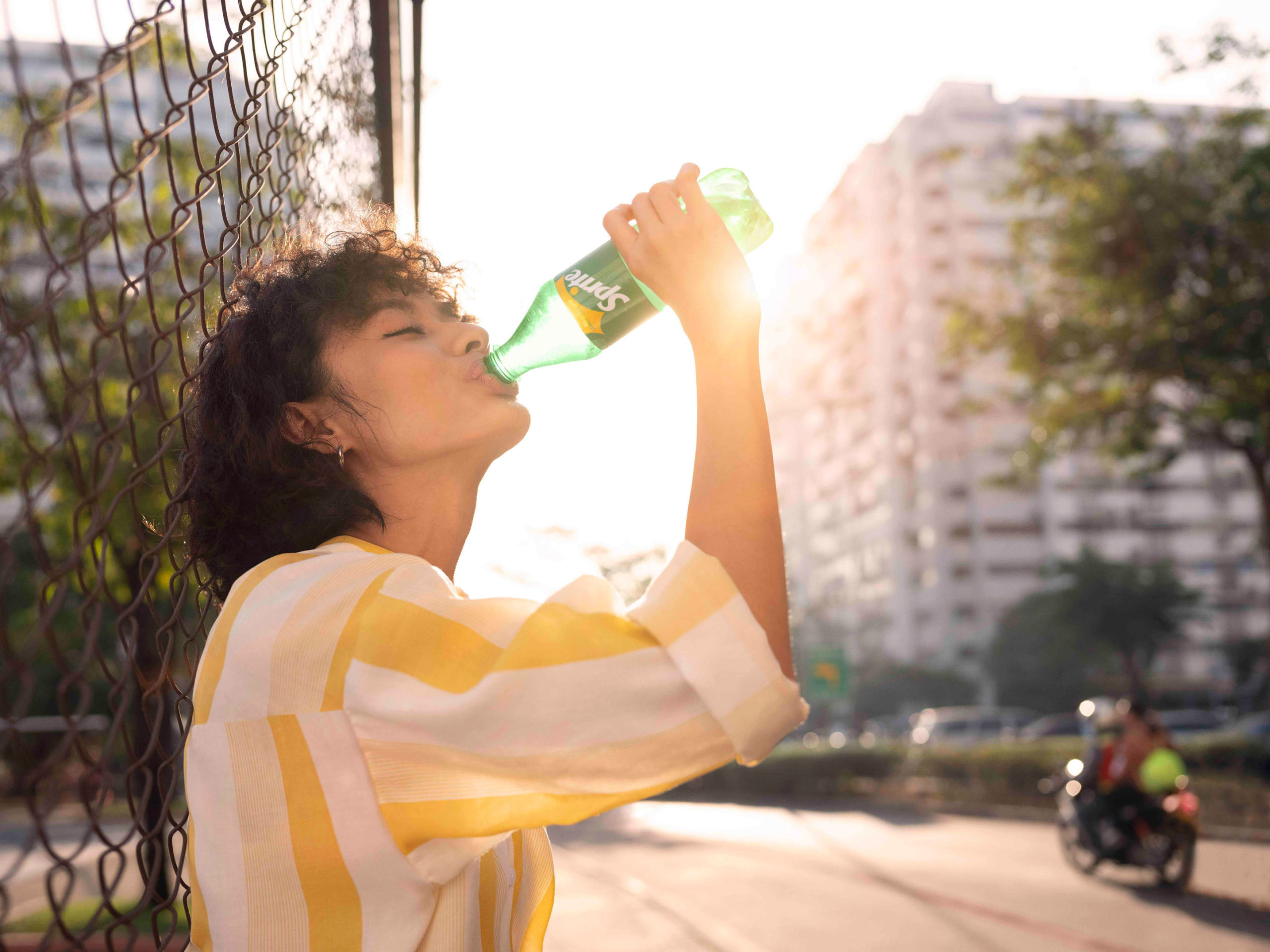 Young adult drinks Sprite from a green bottle outdoors next to a chain-link fence in bright sunlight with blurred urban background.