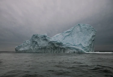 A massive blue and white iceberg floating on a dark ocean under an overcast grey sky, captured in a horizontal photograph by Simon Harsent.