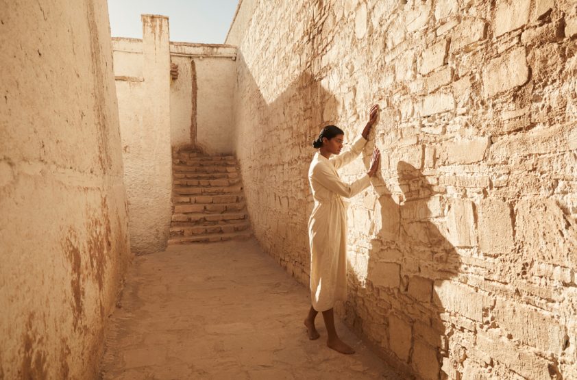 Woman in a cream dress stands barefoot, touching an aged brick wall inside a sunlit desert building with stairs in the background.
