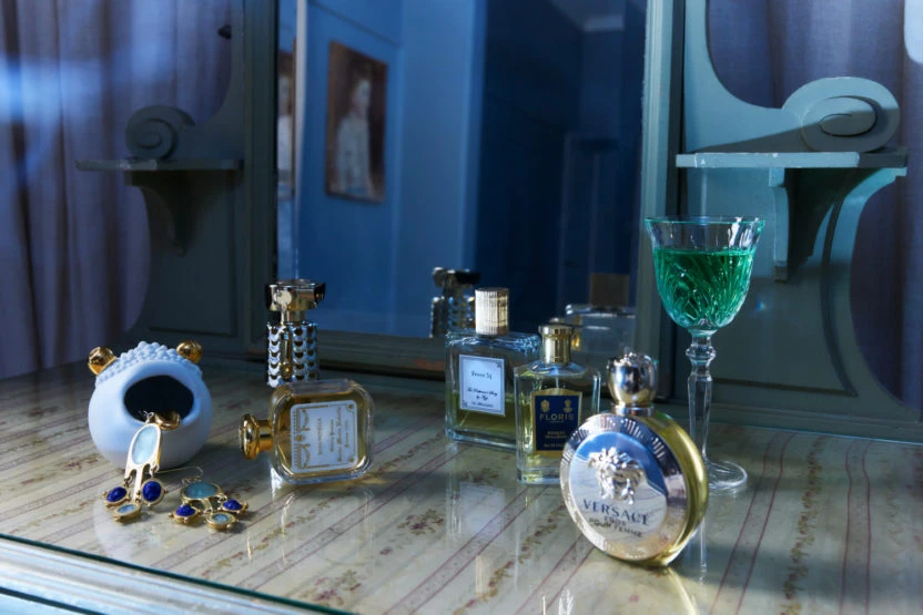 Several perfume bottles, jewelry and a green drink on a vanity table in a dimly lit room with a vintage aesthetic.