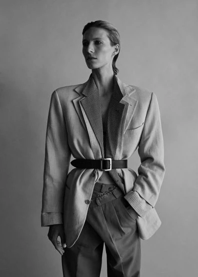 Monochrome portrait of a fashionably dressed adult female with a blazer cinched by a belt, in front of a plain backdrop.