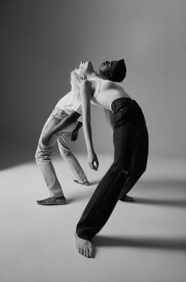 Black and white studio shot of two adult women leaning back to back, one blonde in a white top and jeans, one dark haired in a white top and dark jeans.