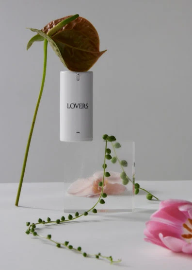 Still life featuring a white cylindrical object labeled 'LOVERS' surrounded by floral elements on a white surface with a gray background.
