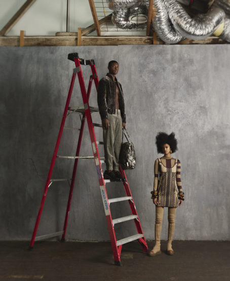 A man stands on a red ladder with a bag next to an adult woman with a large afro in an indoor studio setting.