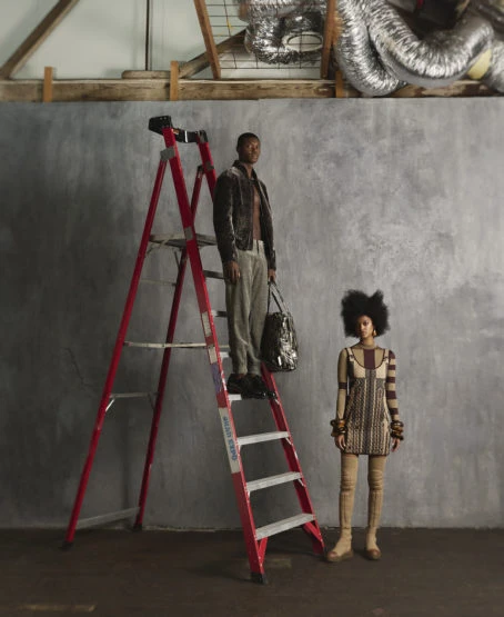 A man stands on a red ladder with a bag next to an adult woman with a large afro in an indoor studio setting.