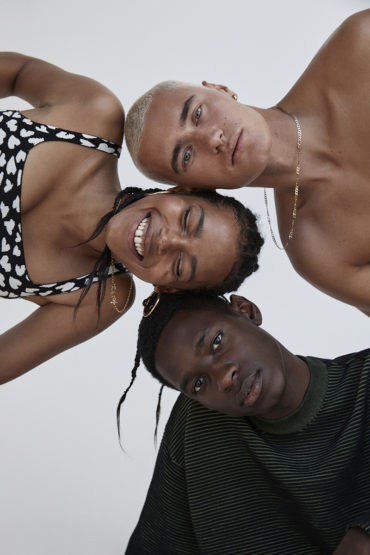 Three young adult friends pose closely together in a bright studio setting. One person wears a black top with white hearts.