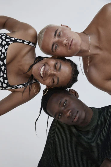 Three young adult friends pose closely together in a bright studio setting. One person wears a black top with white hearts.