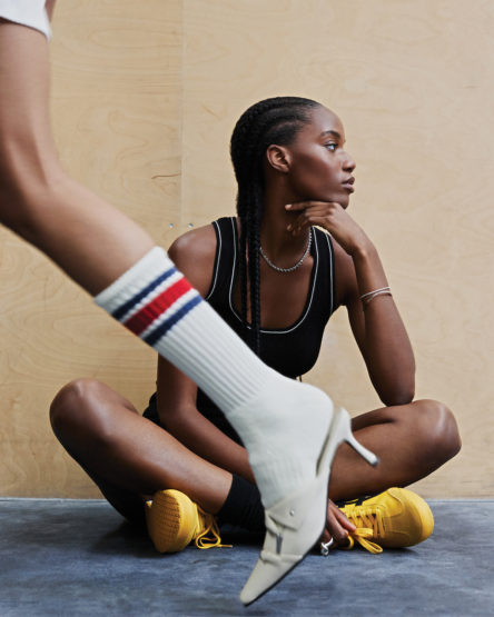 A stylish studio shot featuring a seated Black woman, showcasing fashion accessories, with another person's leg and footwear in focus.