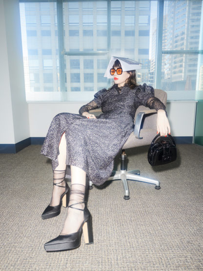 Woman in a dress and heels poses in an office with paper on her head and wearing orange glasses in front of a window.