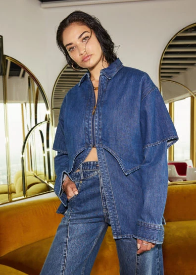 A woman poses indoors wearing a blue denim outfit, featuring a short zipper jacket and wide-legged jeans, against a mustard-colored sofa.