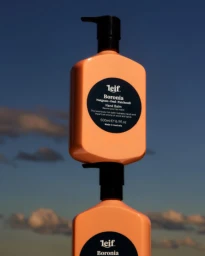 Two orange hand balm bottles stacked vertically against a clear blue sky with soft clouds, photographed by Jedd Cooney