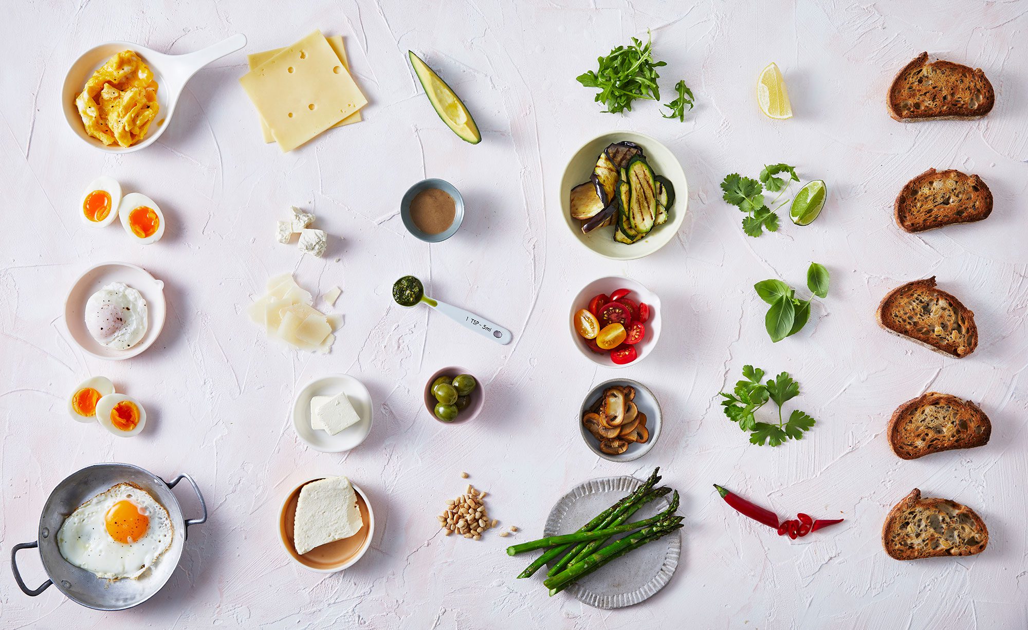 Overhead food flat lay featuring eggs, cheese, vegetables, bread, herbs, and seasoning ingredients arranged on a white textured surface.