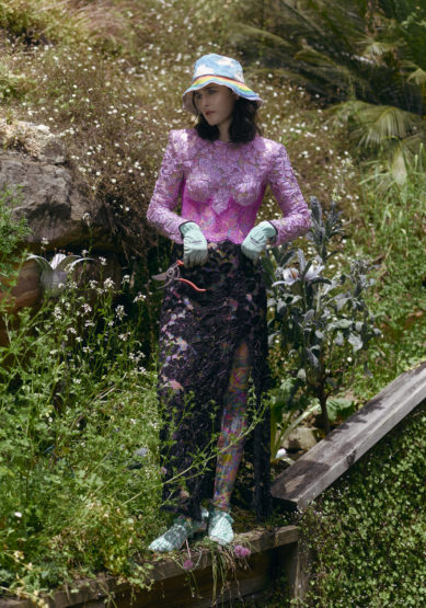 Woman poses in garden wearing pink lace top, black lace skirt, blue bucket hat, and gloves. Gardening shear visible in her hand.