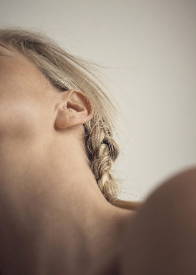 Close-up on the side of a woman's face and neck, featuring blonde hair styled in a braid, and illuminated by diffused, natural light.