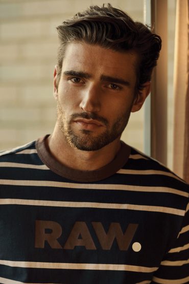 Portrait of an adult male with a beard, wearing a striped shirt with the text RAW, medium shot indoors with natural light.