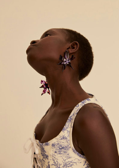 A young Black female model poses with large, jeweled flower earrings and a patterned top against a neutral backdrop in a portrait shot.