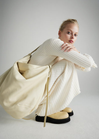 A blonde woman models a slouchy cream-colored shoulder bag and a textured white outfit in a studio setting, capturing a minimalist aesthetic.