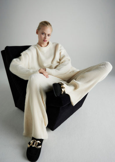 Fashion shot of a woman in a cream sweater and pants, lounging in a chair with decorative flats, against a plain white background.