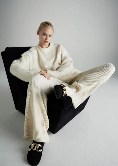 Fashion shot of a woman in a cream sweater and pants, lounging in a chair with decorative flats, against a plain white background.