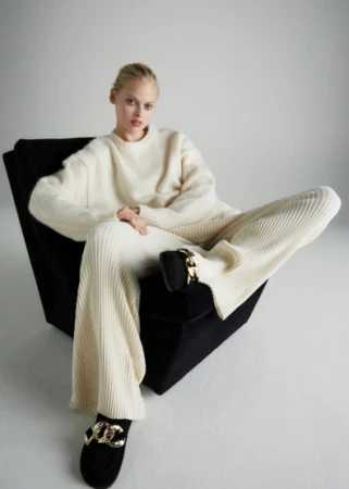 Fashion shot of a woman in a cream sweater and pants, lounging in a chair with decorative flats, against a plain white background.