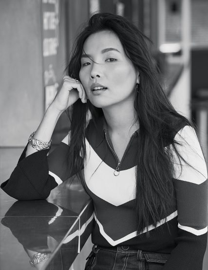 Portrait of a young Asian woman with long hair wearing a patterned sweater, resting her face on her hand, with a contemplative expression.