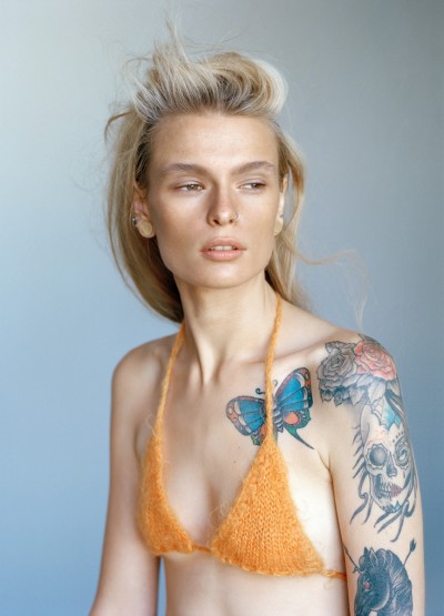 Portrait of a blonde adult female with light skin wearing an orange knit bikini top. Her arm has various tattoos.