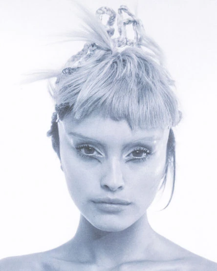Close-up portrait of a fair-skinned woman with light blonde hair styled in an updo, and dramatic makeup, set against a bright white background.