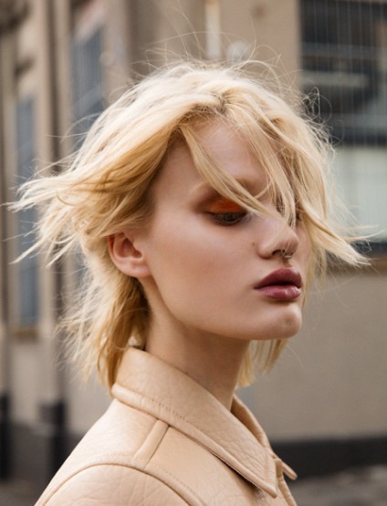 Close-up portrait of a woman with fair skin and blonde hair, wearing a light-colored jacket against a blurred urban backdrop.