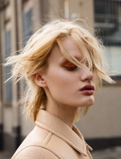 Close-up portrait of a woman with fair skin and blonde hair, wearing a light-colored jacket against a blurred urban backdrop.