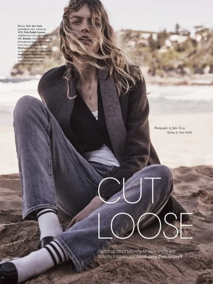 Blonde woman sits on beach with light blue jeans, dark jacket, striped socks, and black loafers. Beach and ocean in the background.