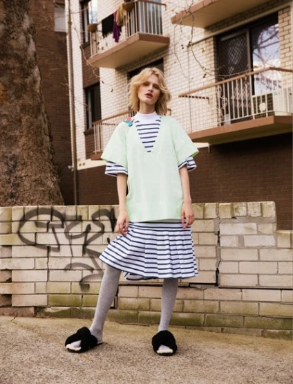 Woman in casual light-green top, striped skirt, and black slippers stands against a weathered brick wall in an urban setting.