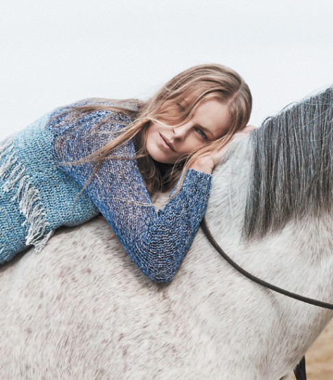 A woman with long blonde hair lies atop a white horse in an outdoor setting, wearing a knitted blue and white sweater, in a lifestyle scene.