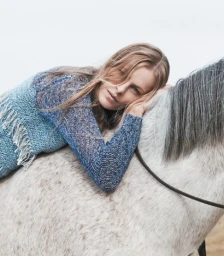 Woman resting her head on a white horse, wearing a textured blue knit top with fringe details against an overcast sky.