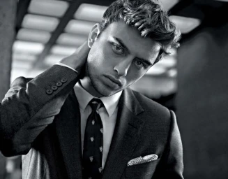 Monochrome portrait of a young man in a dark suit, white shirt, and patterned tie touching the back of his neck
