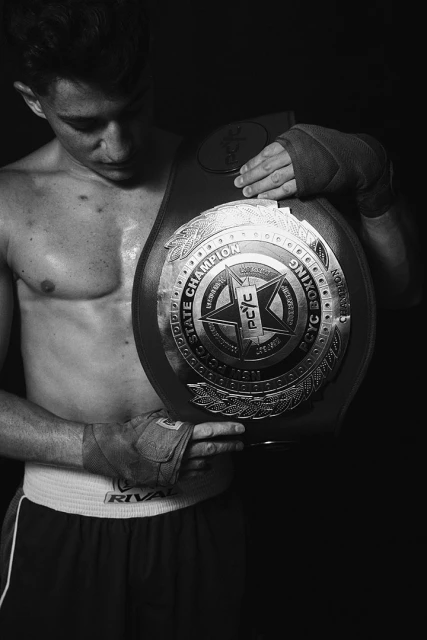 Black and white studio portrait of a young adult male boxer holding a boxing championship belt while looking down. He wears boxing wraps.