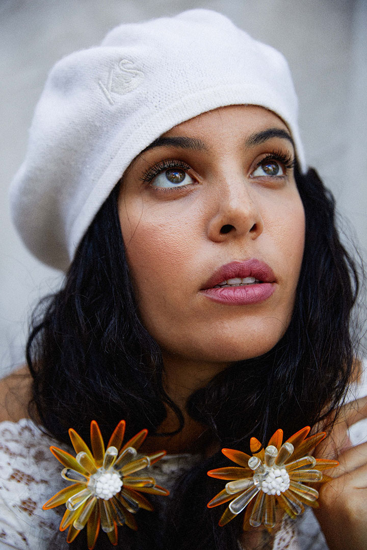 Portrait of a woman with dark curly hair wearing a white beret and statement flower earrings looking up in a close-up studio shot.