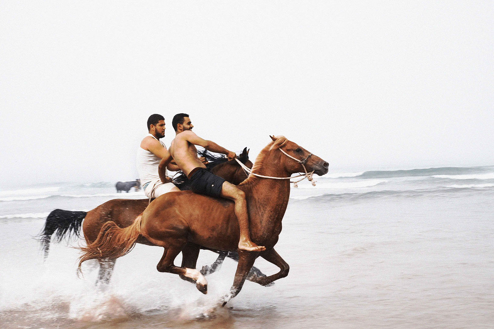 Two men ride chestnut-colored horses through shallow ocean water on a cloudy day, creating splashes around the horses' legs.