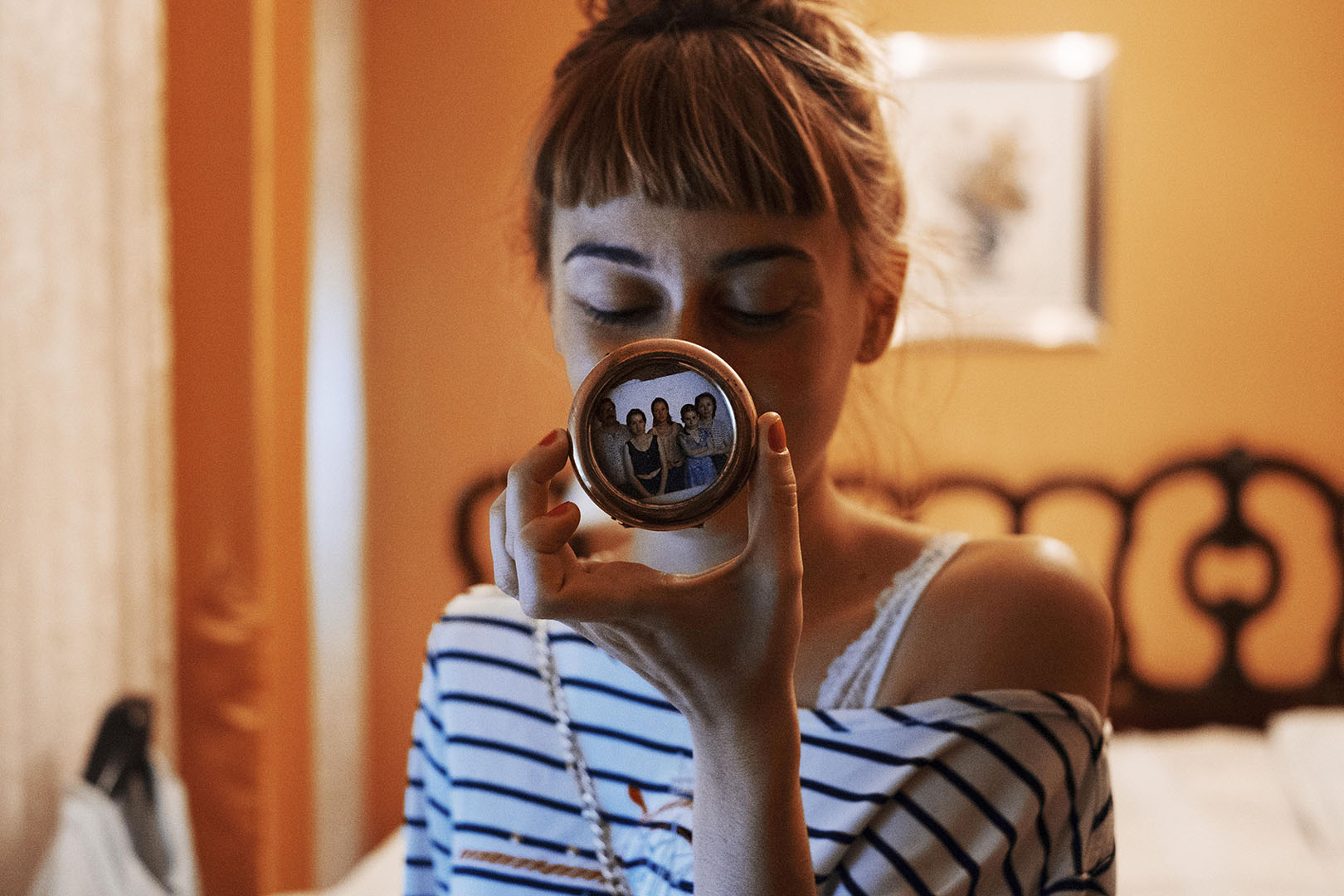 Young woman with bangs holds a round, gold compact mirror reflecting a family photo, indoors with a bed and orange walls in the background.