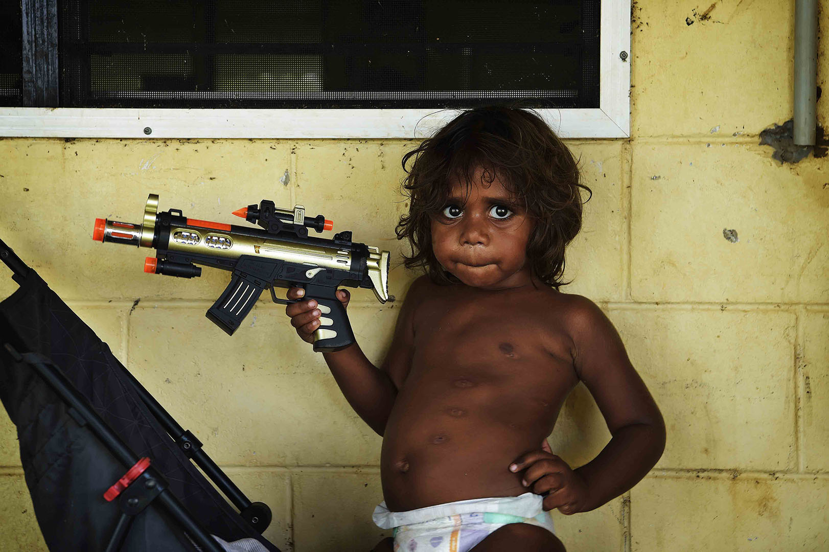 A young child wearing a diaper poses with a toy gun against a weathered wall next to a black stroller inside a room.
