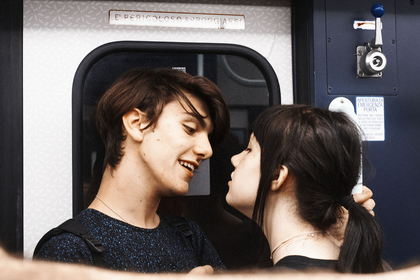 A young couple on a train; the man is smiling and looking at the woman, who is looking up at him in a close-up shot.