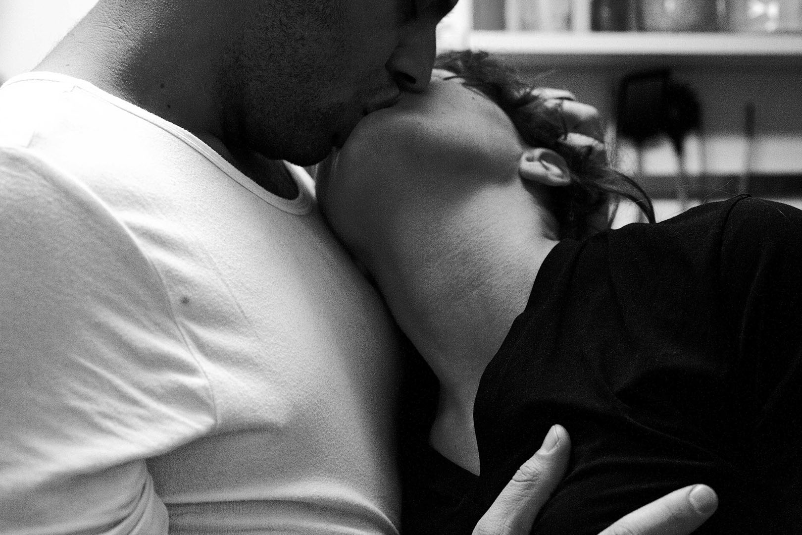 Close-up black and white photograph of two people kissing in a domestic kitchen interior.