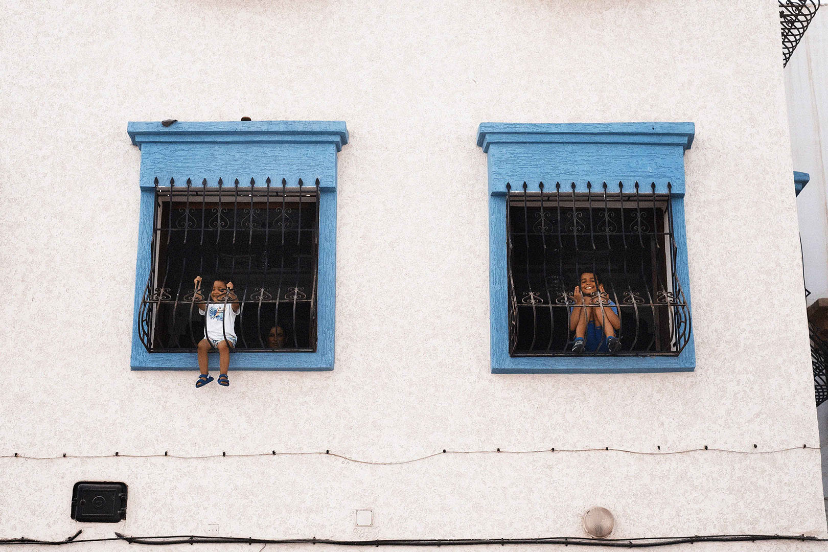 Two young children peer out from windows framed in turquoise on a white building, one clinging to the bars, smiles in the daylight.